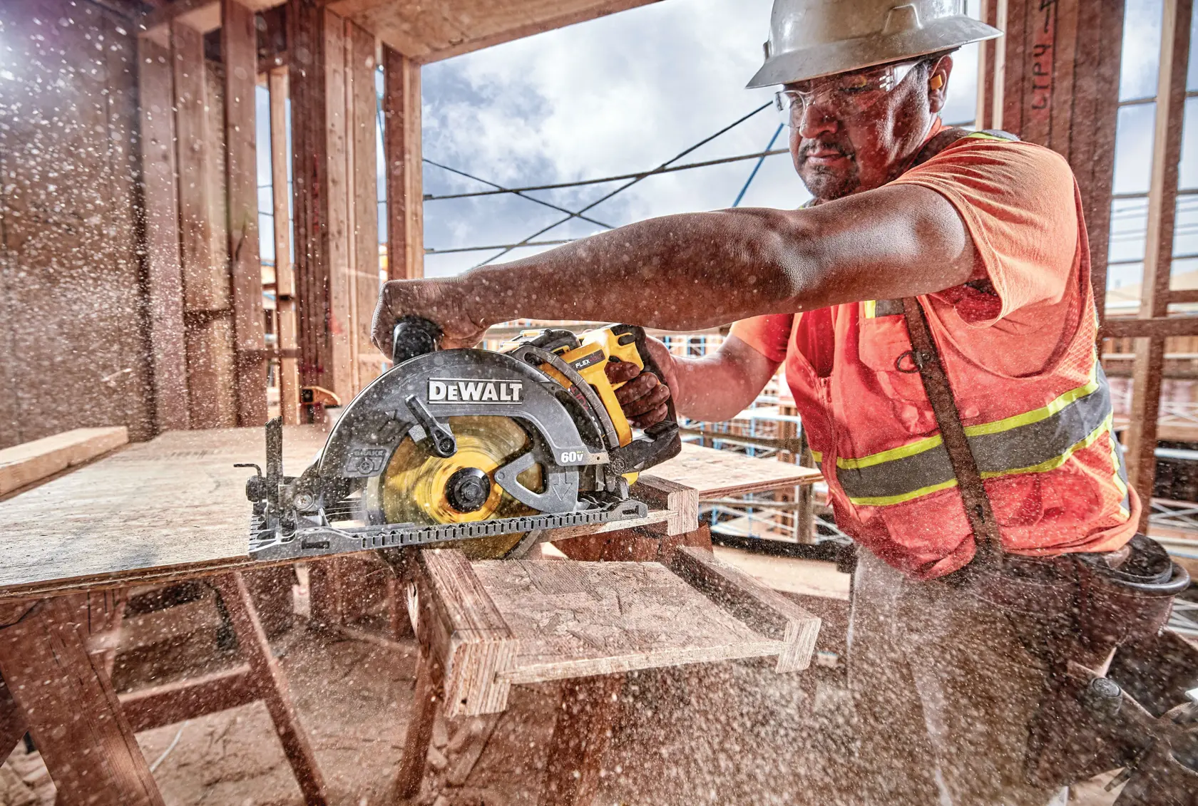 Una persona con equipo de seguridad y un chaleco naranja utiliza una sierra circular DEWALT (modelo DCS577B) para cortar una tabla de madera en una obra de construcción activa. El aserrín vuela por el aire, destacando la potencia de la herramienta.