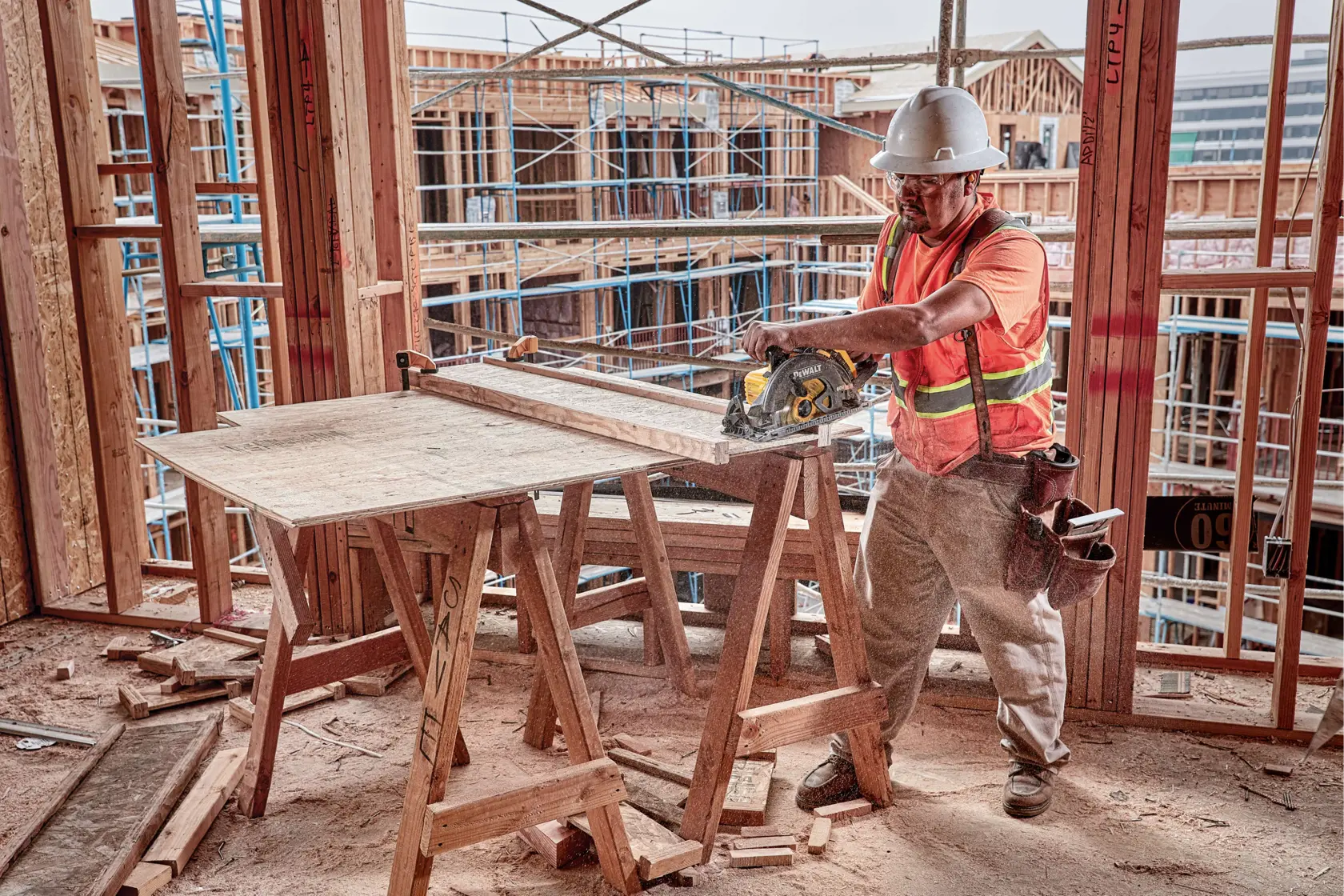 Un trabajador de la construcción con casco y chaleco de seguridad utiliza una sierra circular DEWALT DCS577B para cortar madera en una obra. El trabajador está rodeado de materiales de construcción y andamios y trabaja en una mesa improvisada de madera.