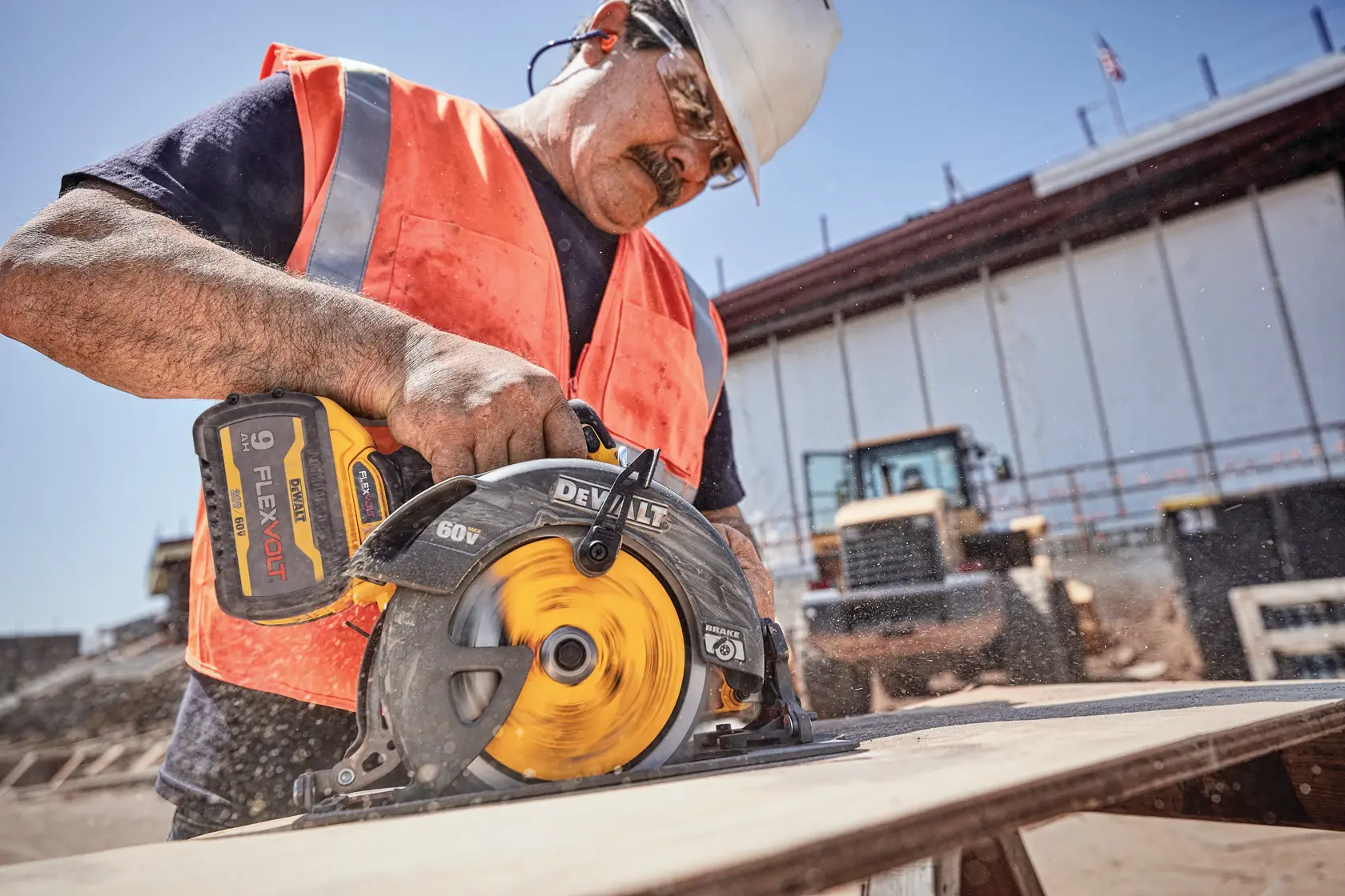 Un trabajador de construcción con chaleco y casco de seguridad está usando una sierra circular inalámbrica DEWALT 60V FLEXVOLT para cortar una tabla de madera en un sitio de construcción. El rostro del trabajador está difuminado por motivos de privacidad.