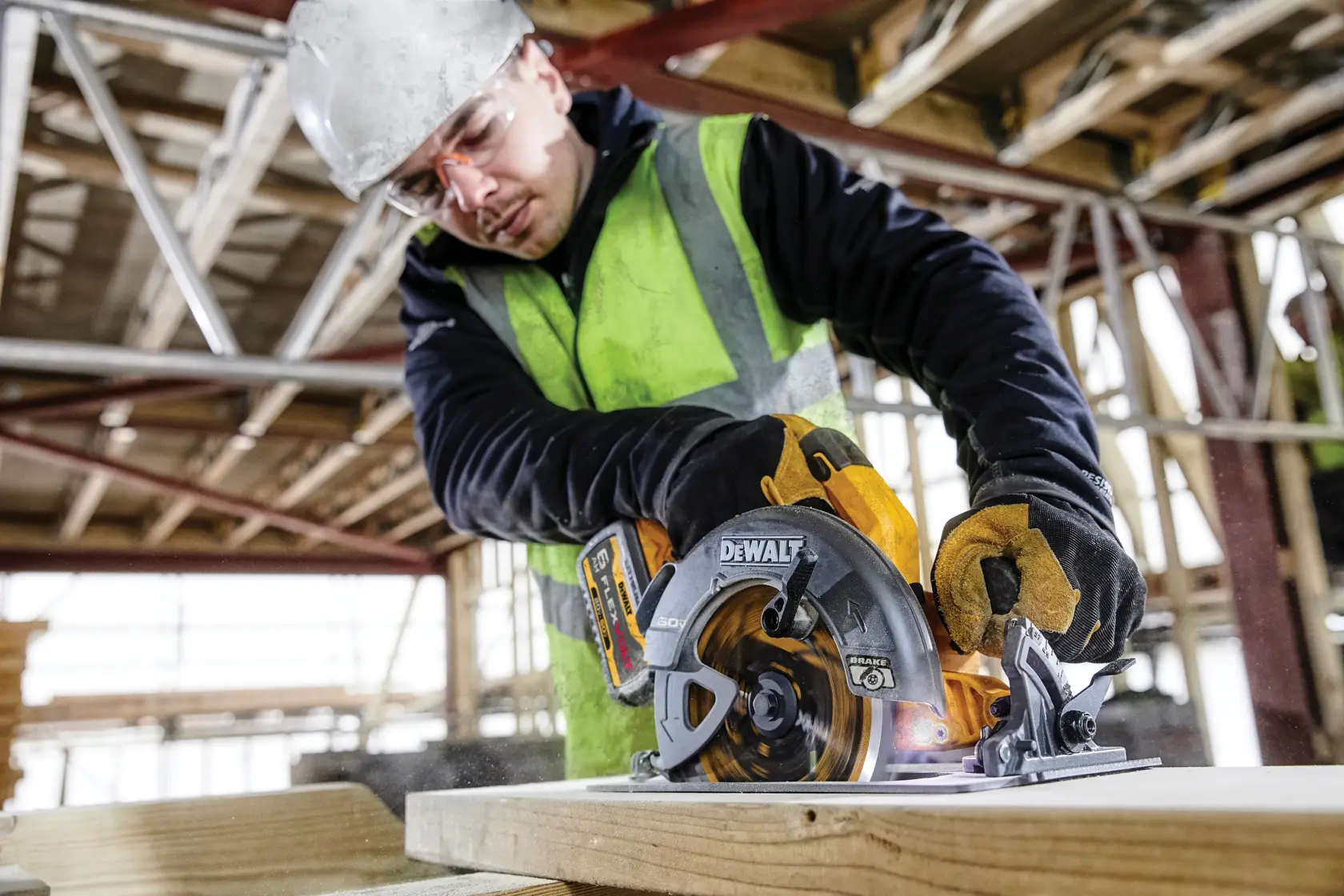 Un trabajador con chaleco de alta visibilidad y guantes utiliza una sierra circular inalámbrica DEWALT para cortar madera en un sitio de construcción. El rostro de la persona está desenfocado para garantizar su privacidad.