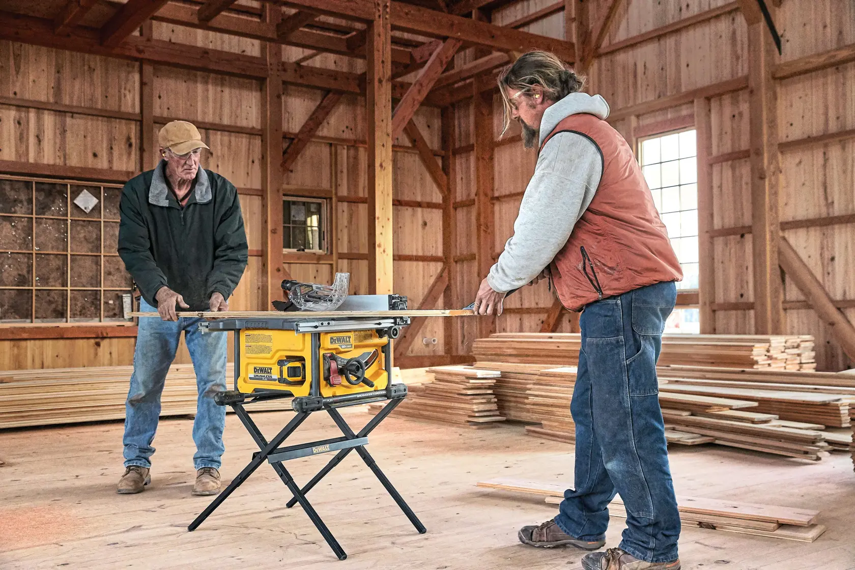 Dos personas trabajan juntas en un taller de madera, utilizando una sierra de mesa amarilla DEWALT colocada sobre un soporte para cortar una pieza de madera. Al fondo se ven pilas de madera y paredes de madera, con luz natural entrando por las ventanas.