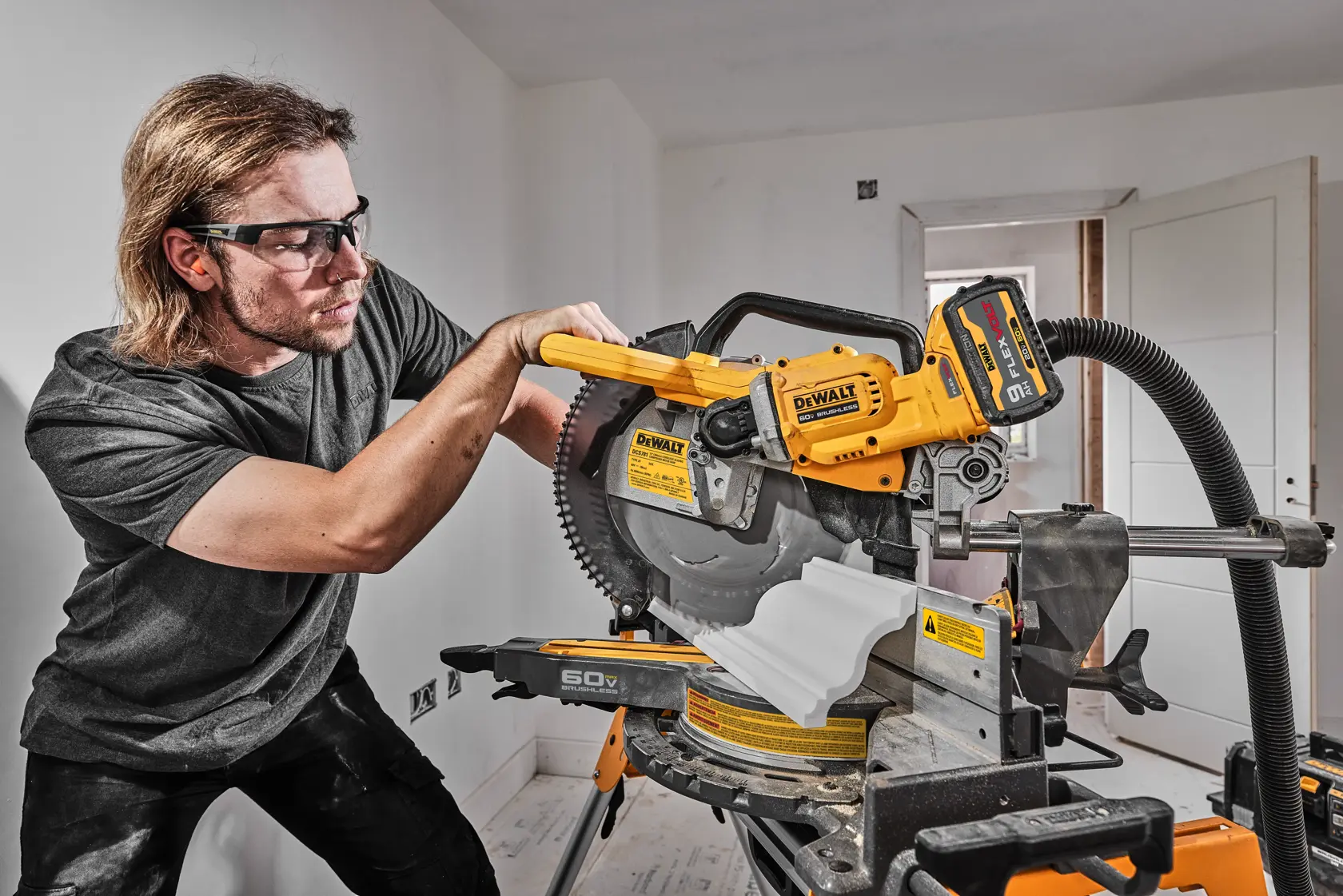 Una persona está utilizando una sierra ingletadora DEWALT DCS781B para cortar una moldura blanca en un taller o entorno de construcción. La sierra es de color amarillo y negro y tiene una manguera de extracción de polvo conectada.