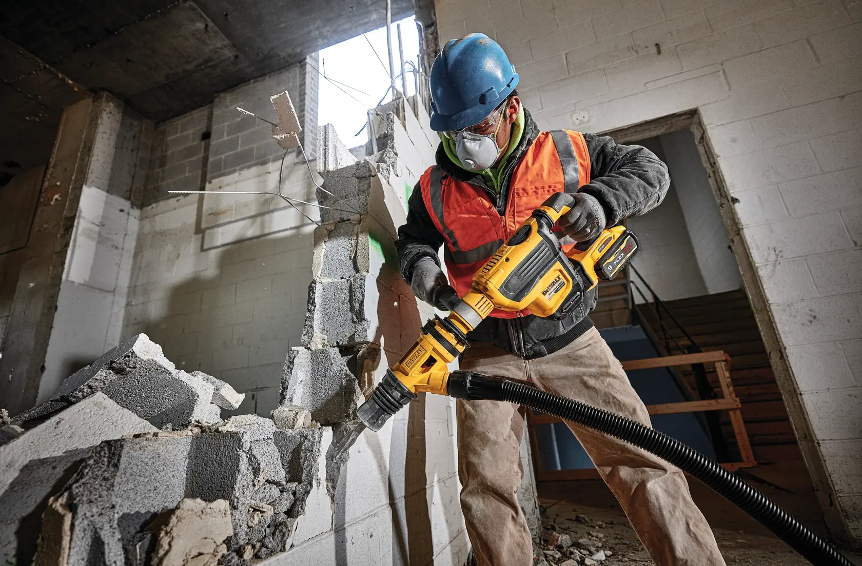 Un trabajador de la construcción con casco azul y chaleco de seguridad naranja utiliza una herramienta eléctrica amarilla DEWALT para demoler una pared de bloques de cemento dentro de un edificio en construcción. Los escombros están dispersos por la zona.