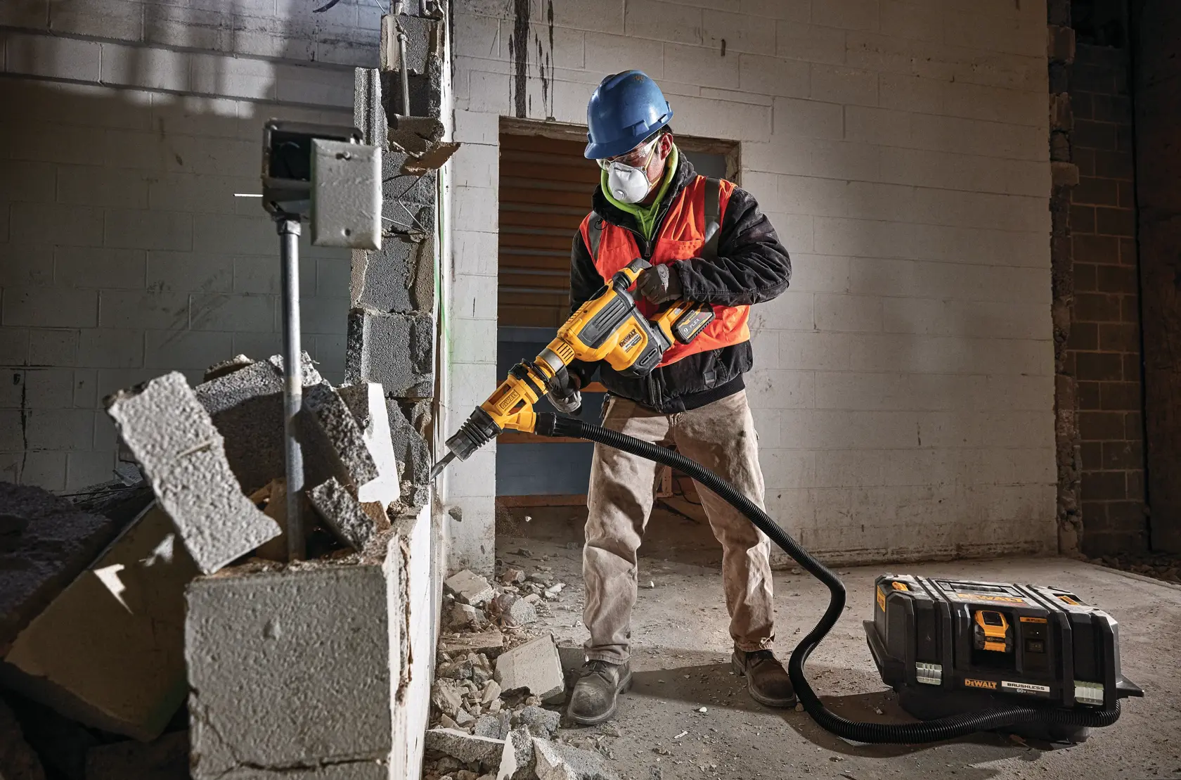 Un trabajador de la construcción con casco azul y chaleco de seguridad naranja opera un extractor de polvo DEWALT DCV585B conectado a una herramienta de demolición para romper bloques de cemento dentro de un edificio.