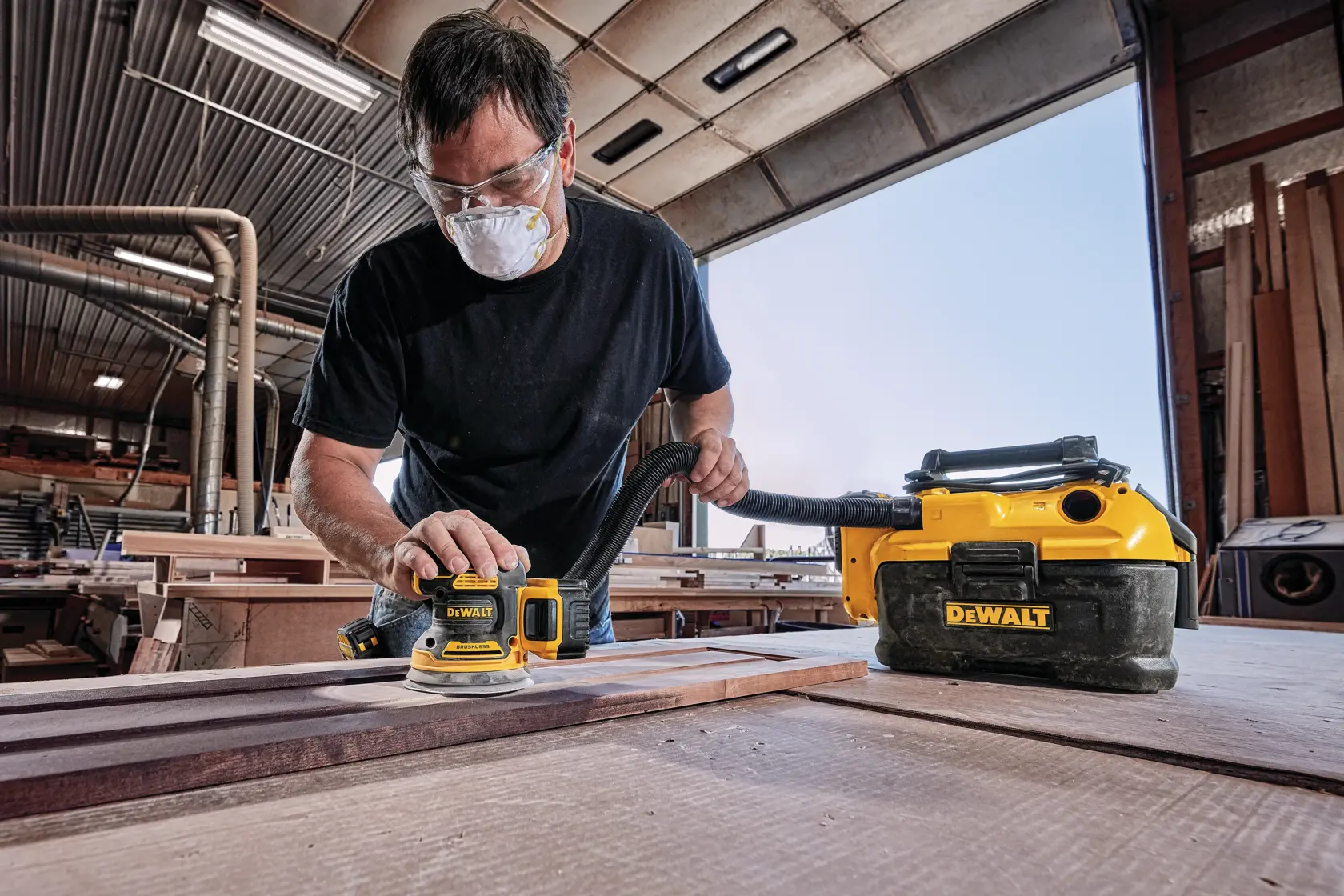 Una persona usando una lijadora inalámbrica DEWALT DCW210B en un taller de carpintería, con una aspiradora DEWALT conectada para la extracción de polvo. El rostro de la persona está difuminado por privacidad.