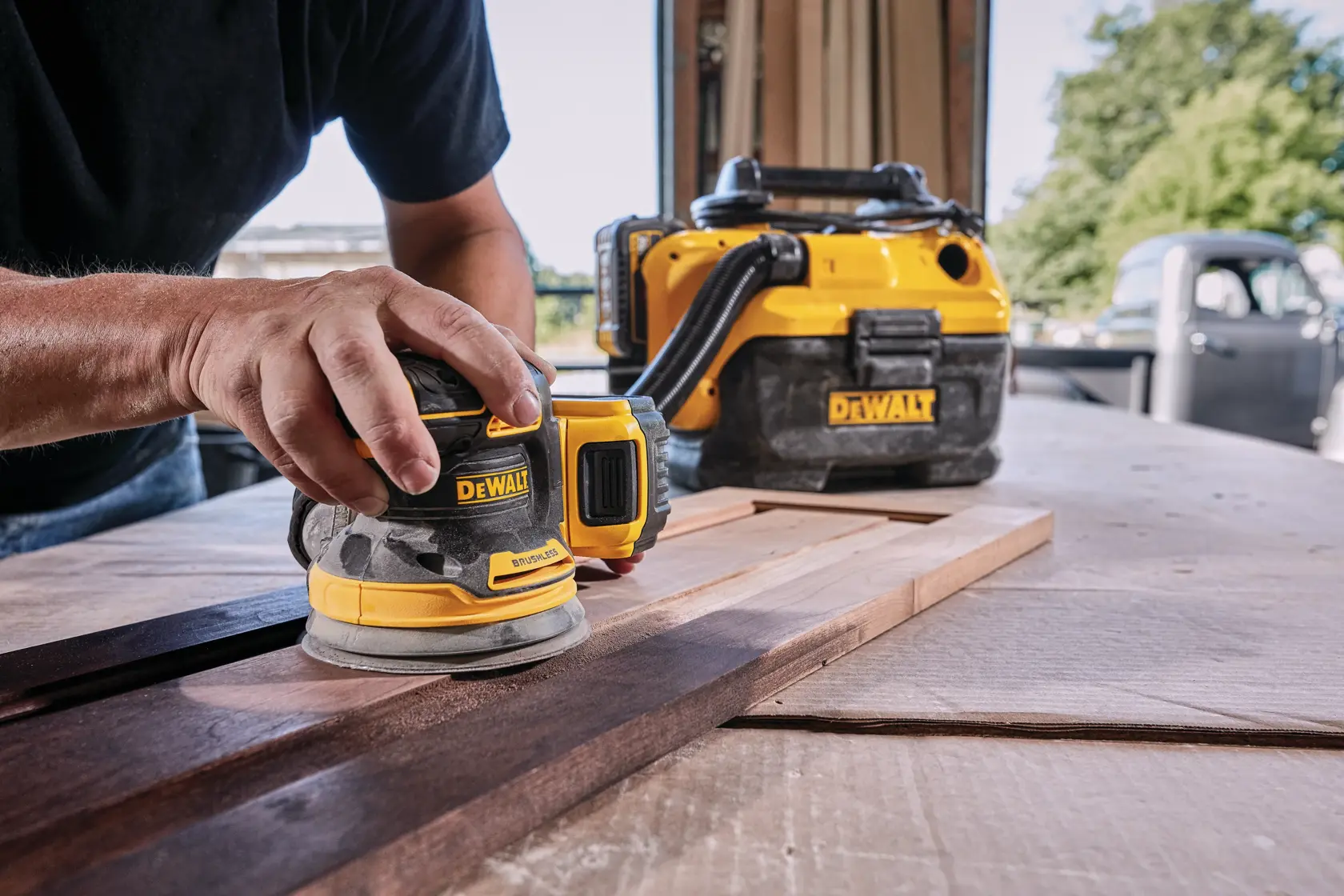 Una persona utiliza una lijadora orbital inalámbrica DEWALT sobre una tabla de madera, con un aspirador DEWALT al fondo sobre un banco de trabajo en un taller.