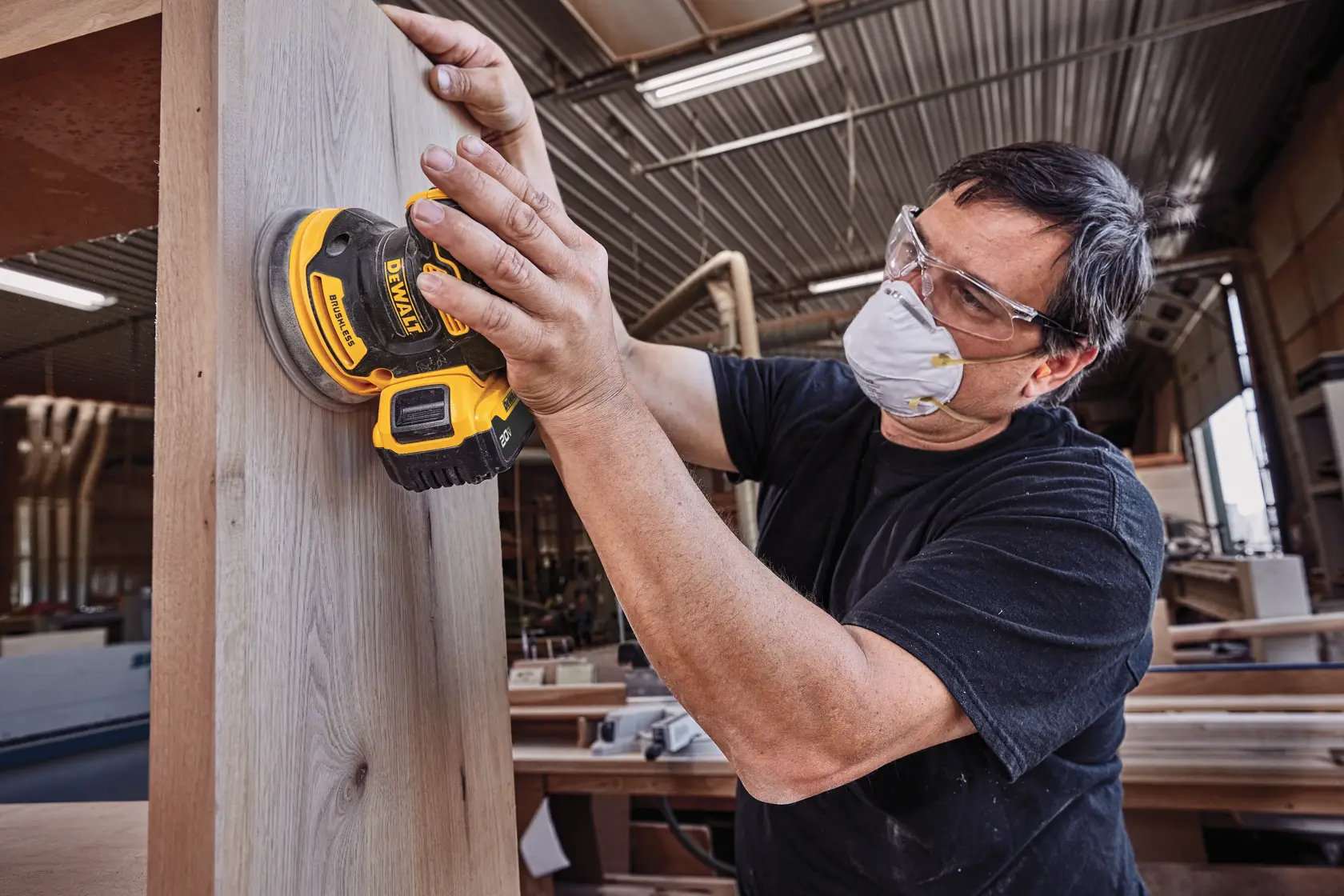 Una persona con mascarilla para polvo está usando una lijadora inalámbrica DEWALT (modelo DCW210B) para lijar una superficie de madera en un taller.
