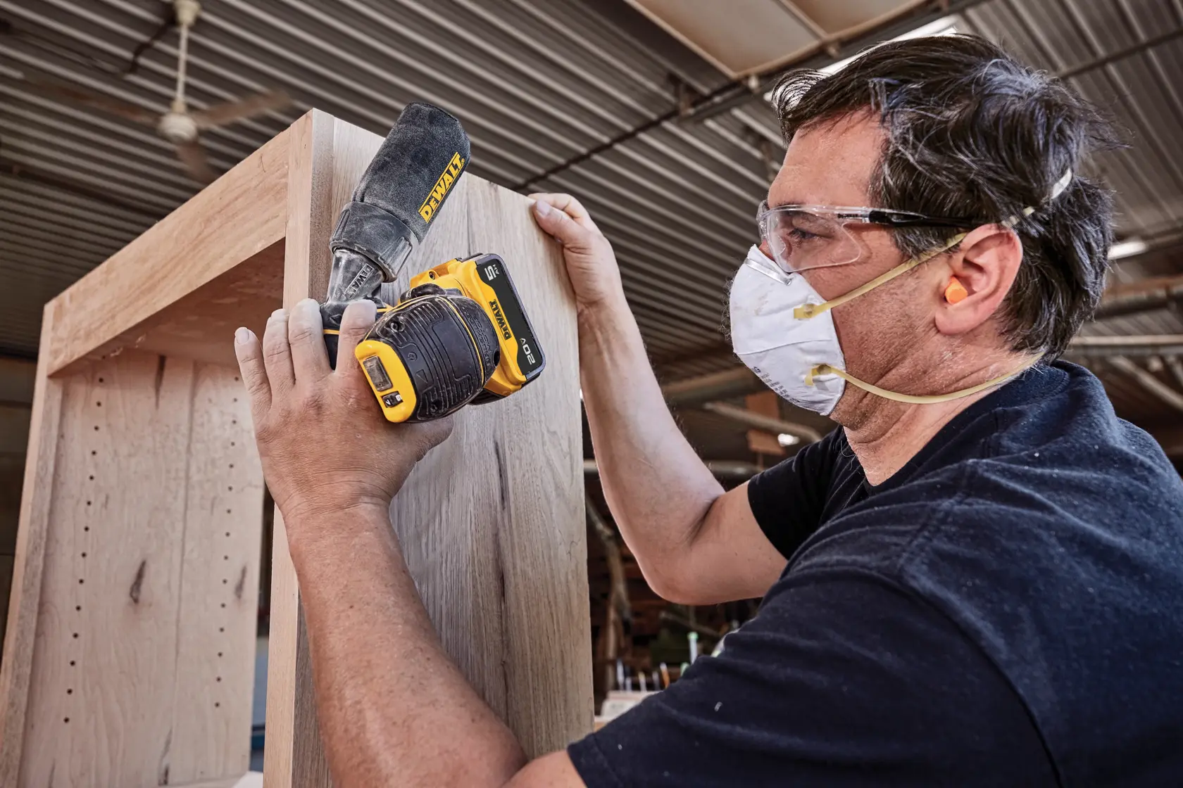 Una persona con protección auditiva y mascarilla para polvo utiliza una lijadora inalámbrica DEWALT para suavizar la superficie de un gabinete de madera en un taller.