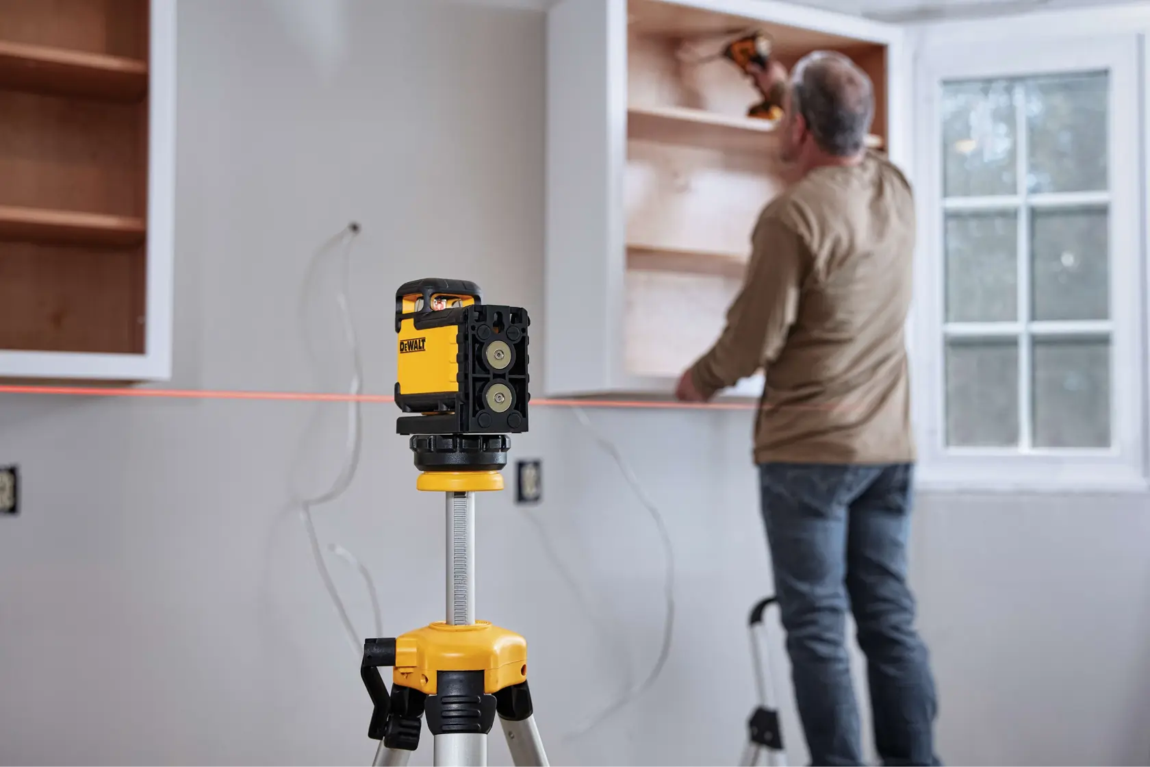 Un nivel láser DEWALT montado en un trípode proyecta una línea roja sobre una pared, ayudando a una persona a instalar gabinetes en una habitación con ventana.