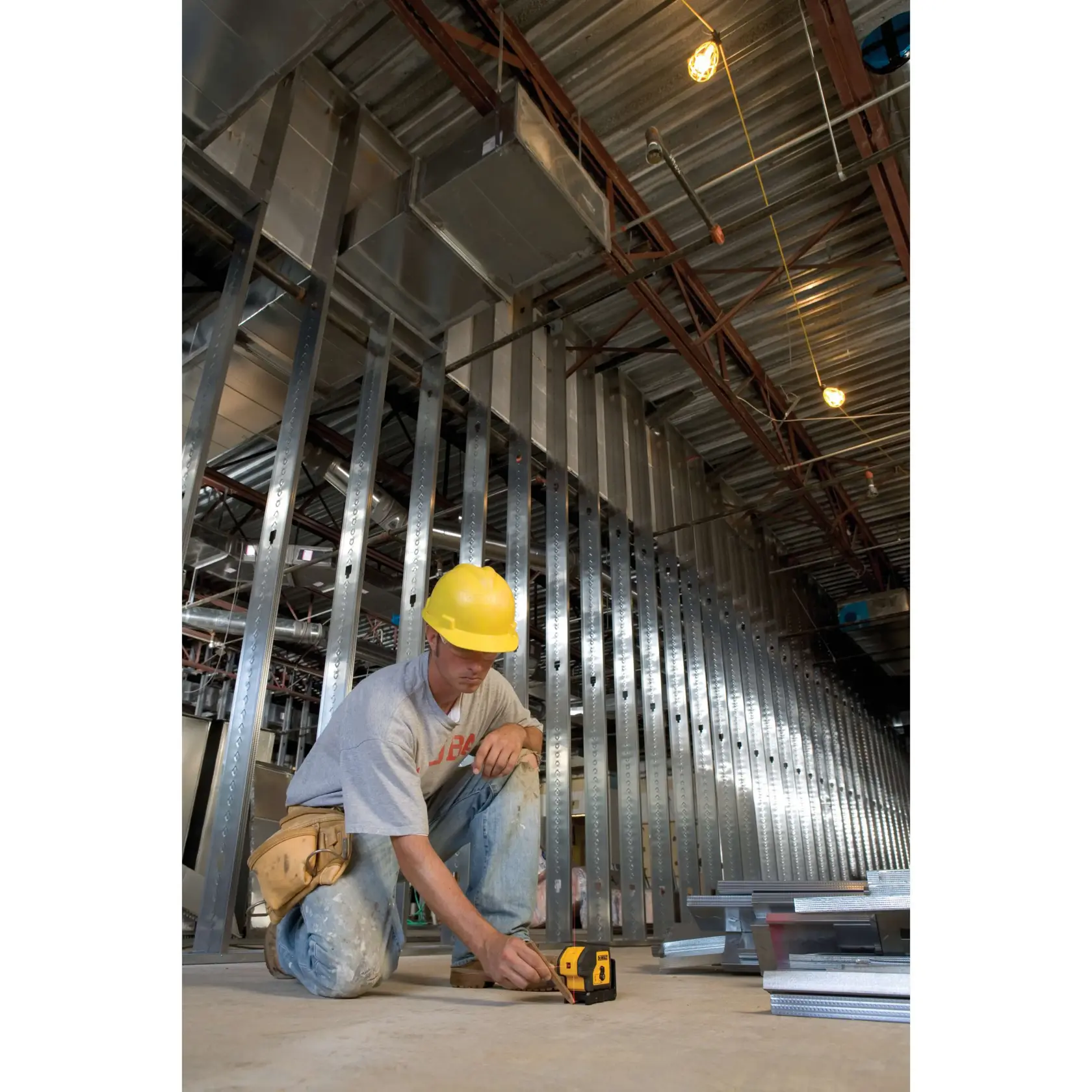 Un trabajador de la construcción con casco amarillo está arrodillado sobre el suelo de un edificio en construcción. El trabajador está utilizando un nivel láser amarillo y negro DEWALT (modelo DW083K) en el suelo de concreto, rodeado de montantes metálicos y materiales de construcción.