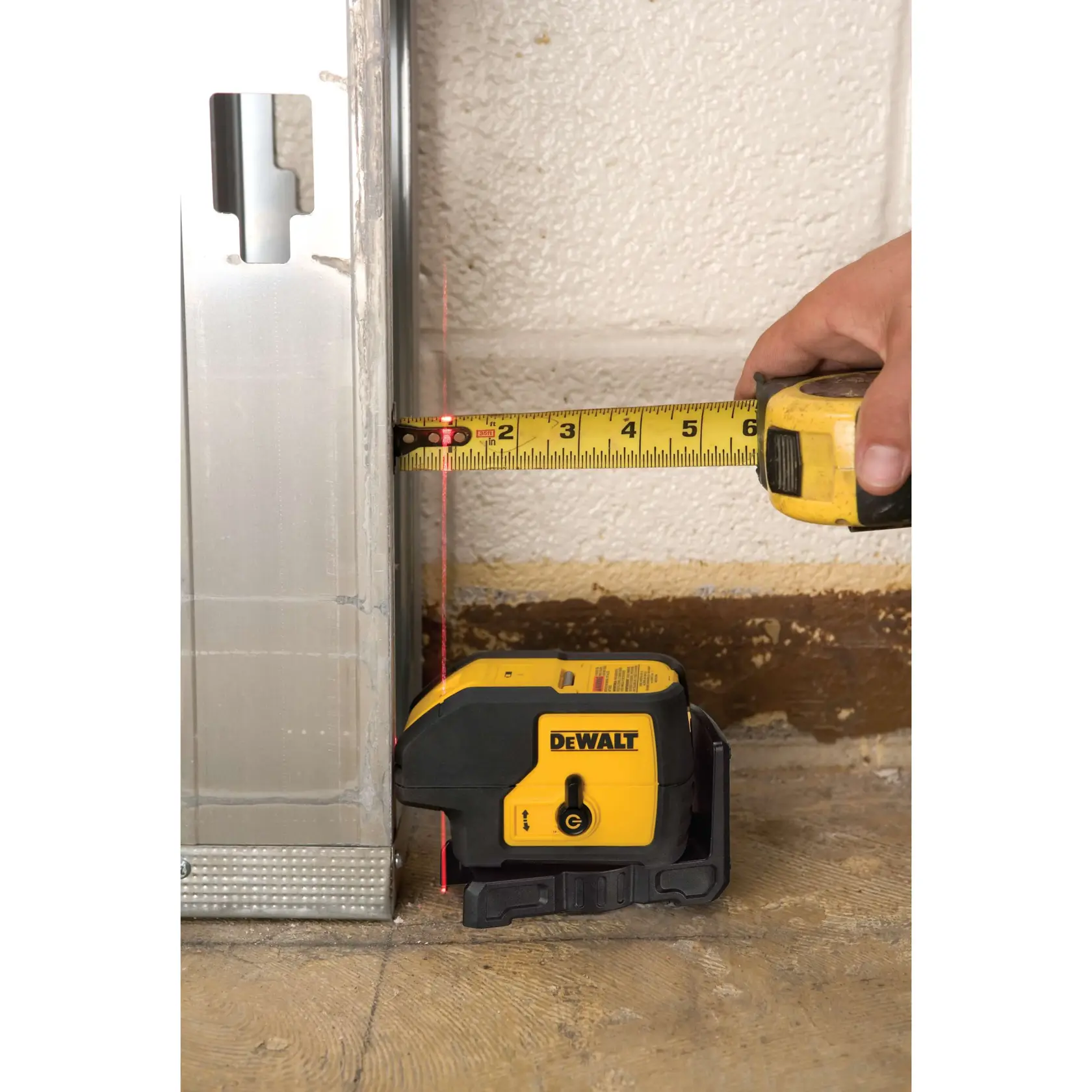 Nivel láser DEWALT alinea marco metálico con pared, cinta métrica muestra la distancia.