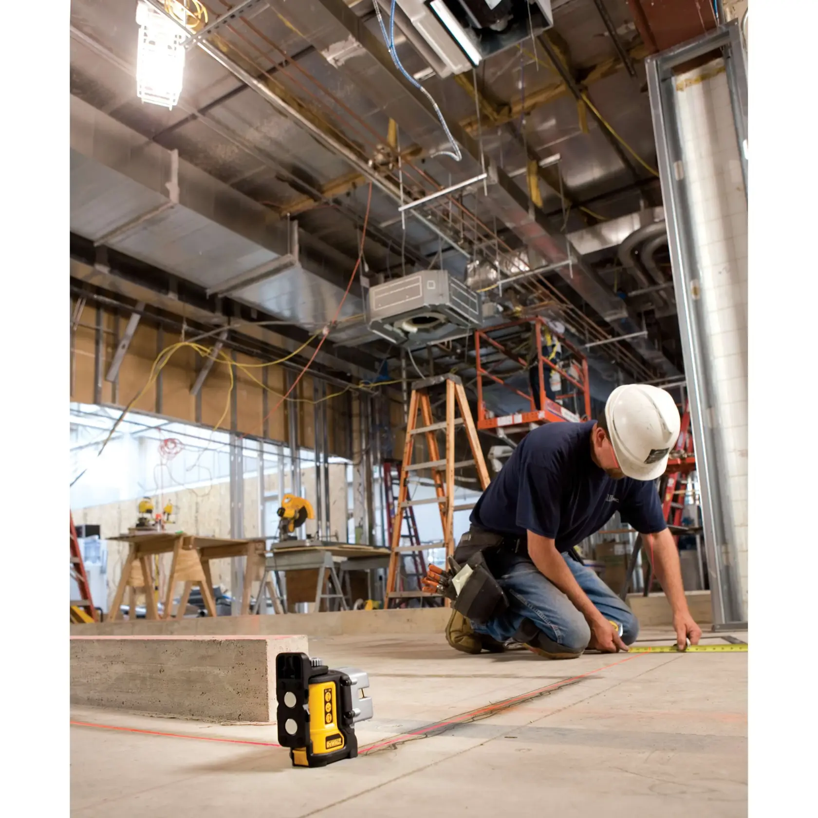 Un nivel láser DEWALT DW089K está colocado en el suelo en un sitio de construcción mientras un trabajador con casco y cinturón de herramientas mide con una cinta métrica en el piso. Al fondo se observa equipo de construcción, escaleras y tuberías expuestas en el techo.