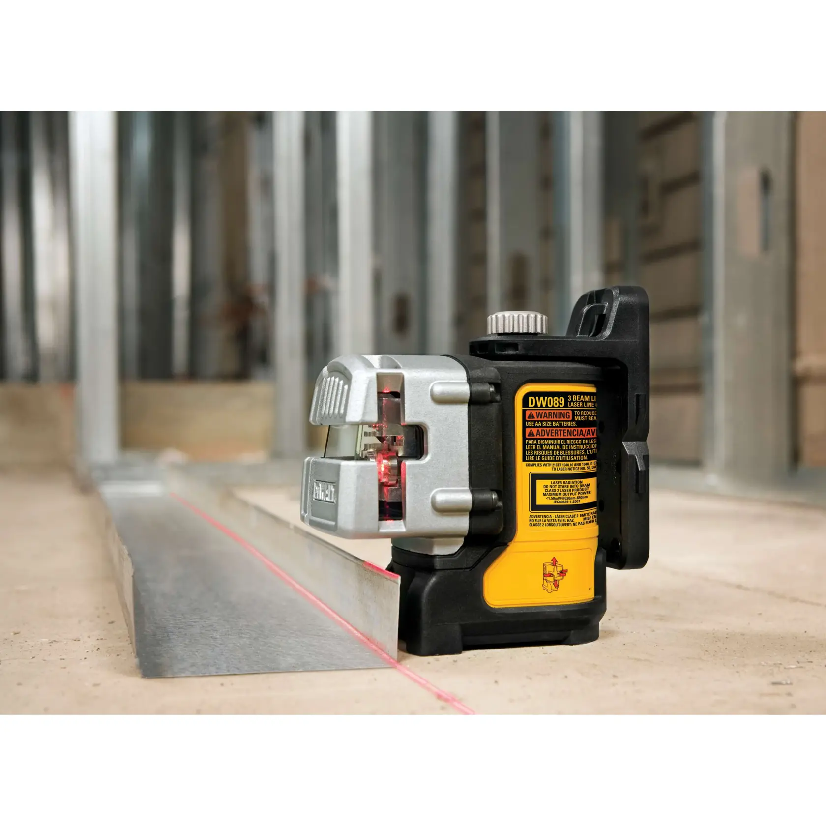 Nivel láser de tres haces DEWALT DW089K situado en el suelo de una obra, proyectando una línea láser roja a lo largo de una guía de estructura metálica con montantes de pared de metal al fondo.