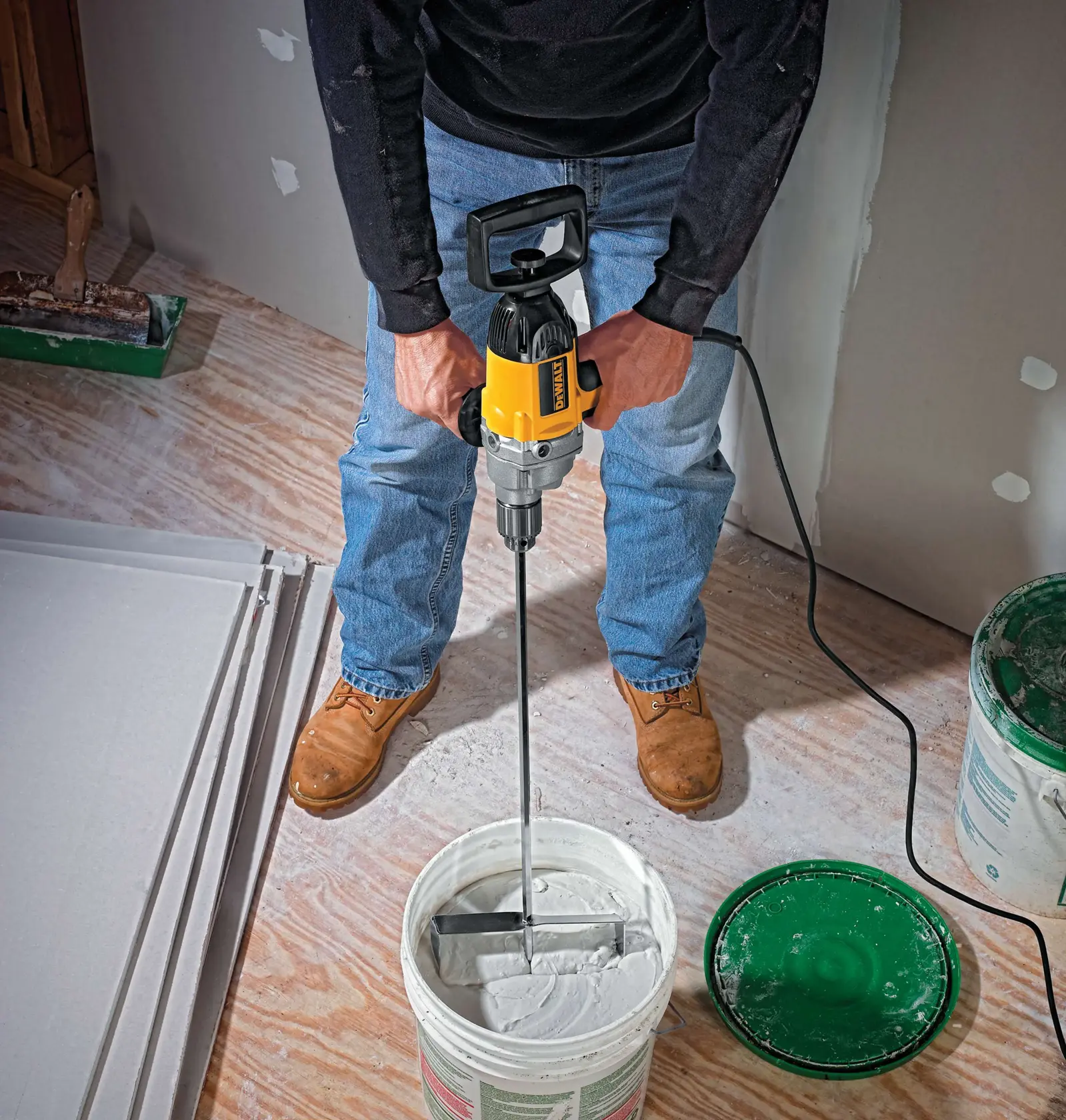Una persona utiliza un taladro eléctrico DEWALT DW130V, equipado con una paleta mezcladora, para revolver un compuesto blanco dentro de un balde. La escena está situada sobre un piso de madera con láminas de yeso y otros materiales de construcción visibles.