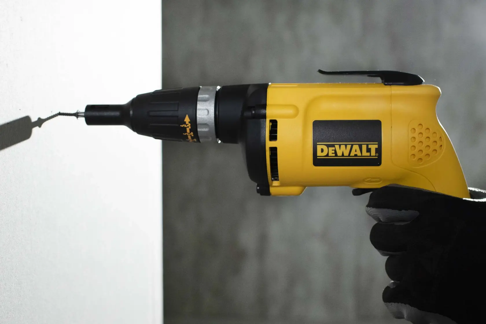 Un destornillador eléctrico amarillo DEWALT está siendo usado para atornillar en una pared. La herramienta está sostenida por una mano con guante.