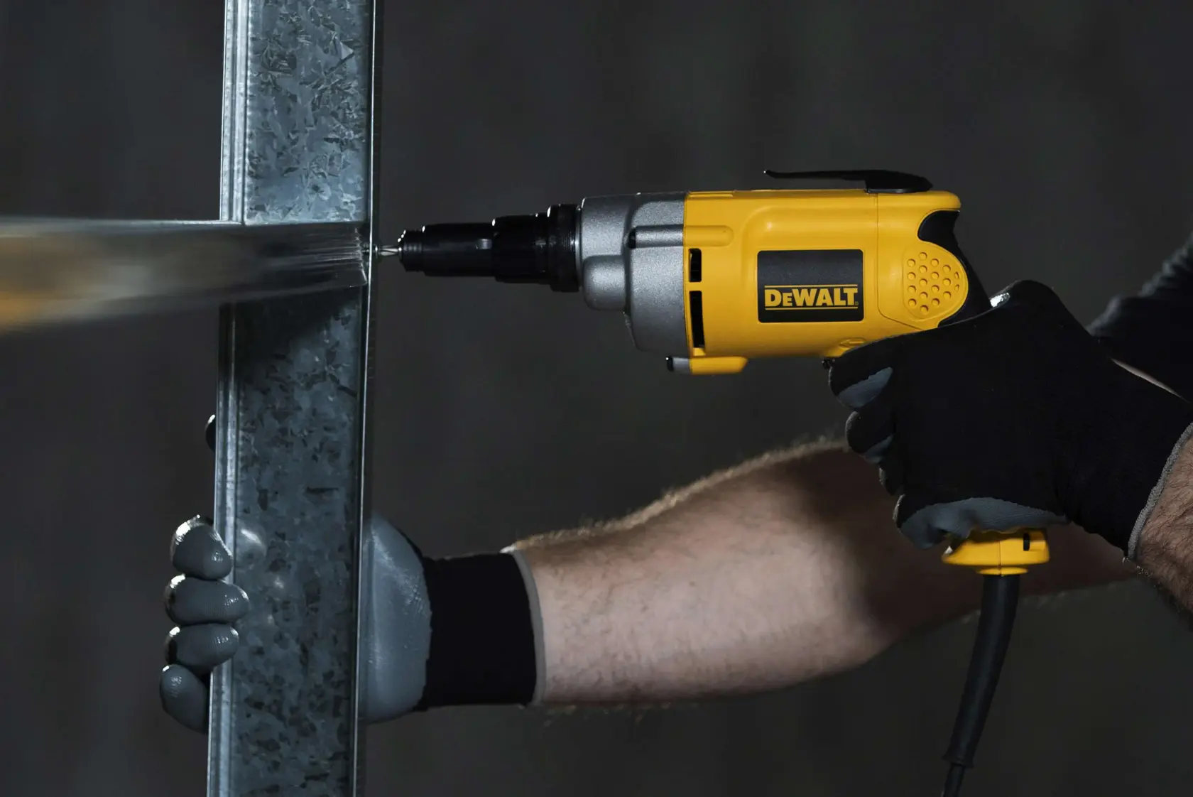 Una persona con guantes utiliza un destornillador eléctrico amarillo y negro DEWALT DW268 para perforar un marco metálico.