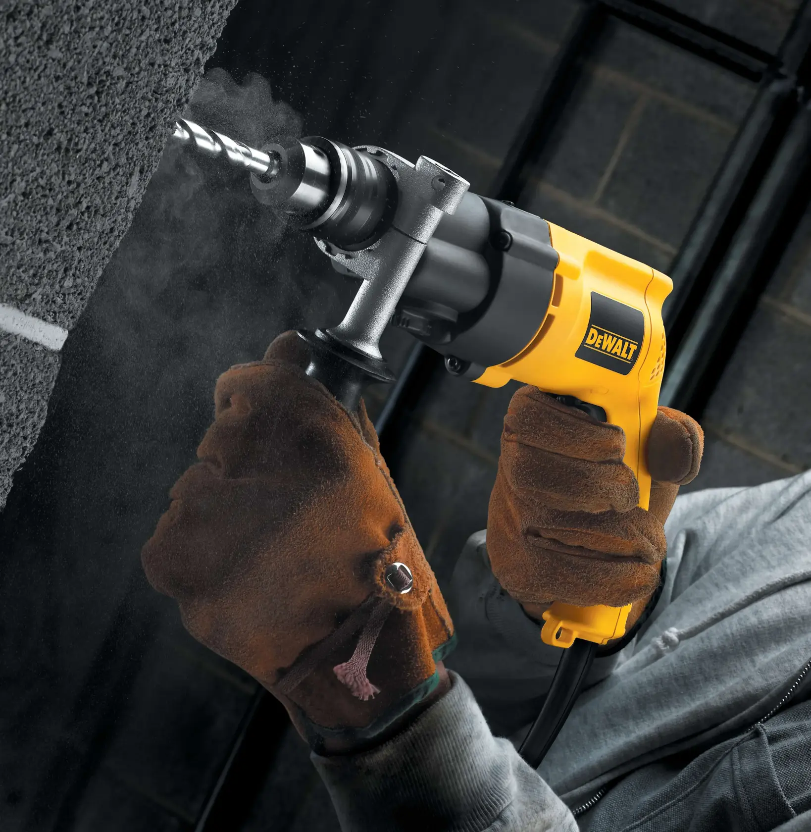 Una persona con guantes de trabajo marrones utiliza un taladro eléctrico amarillo DEWALT para perforar una pared de hormigón; se ve polvo alrededor de la broca.