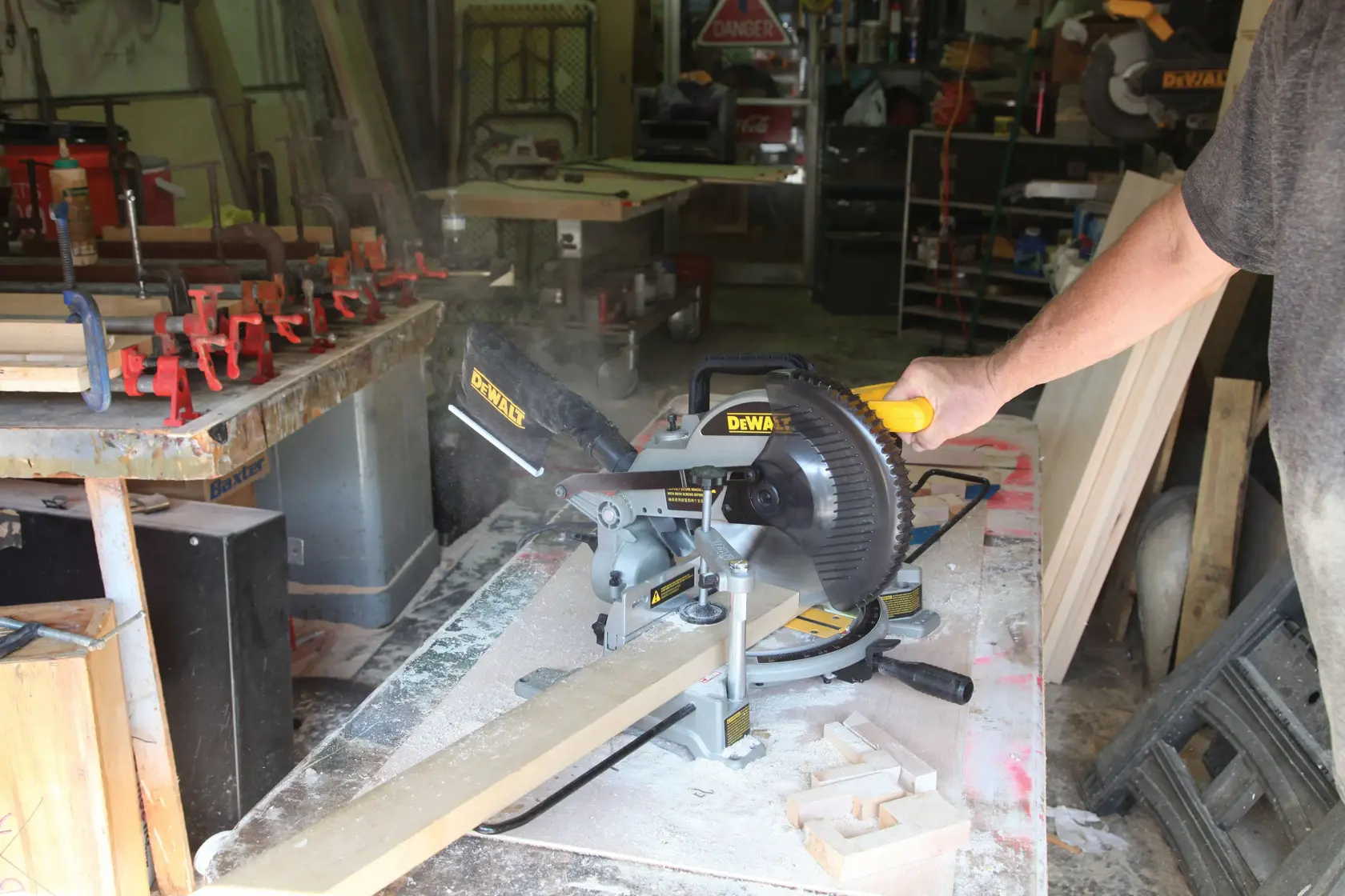 Una persona opera una sierra de inglete DEWALT (SKU: DW714) para cortar una tabla de madera en un taller. Hay polvo de madera y abrazaderas visibles en la mesa de trabajo, con varias herramientas y materiales al fondo.