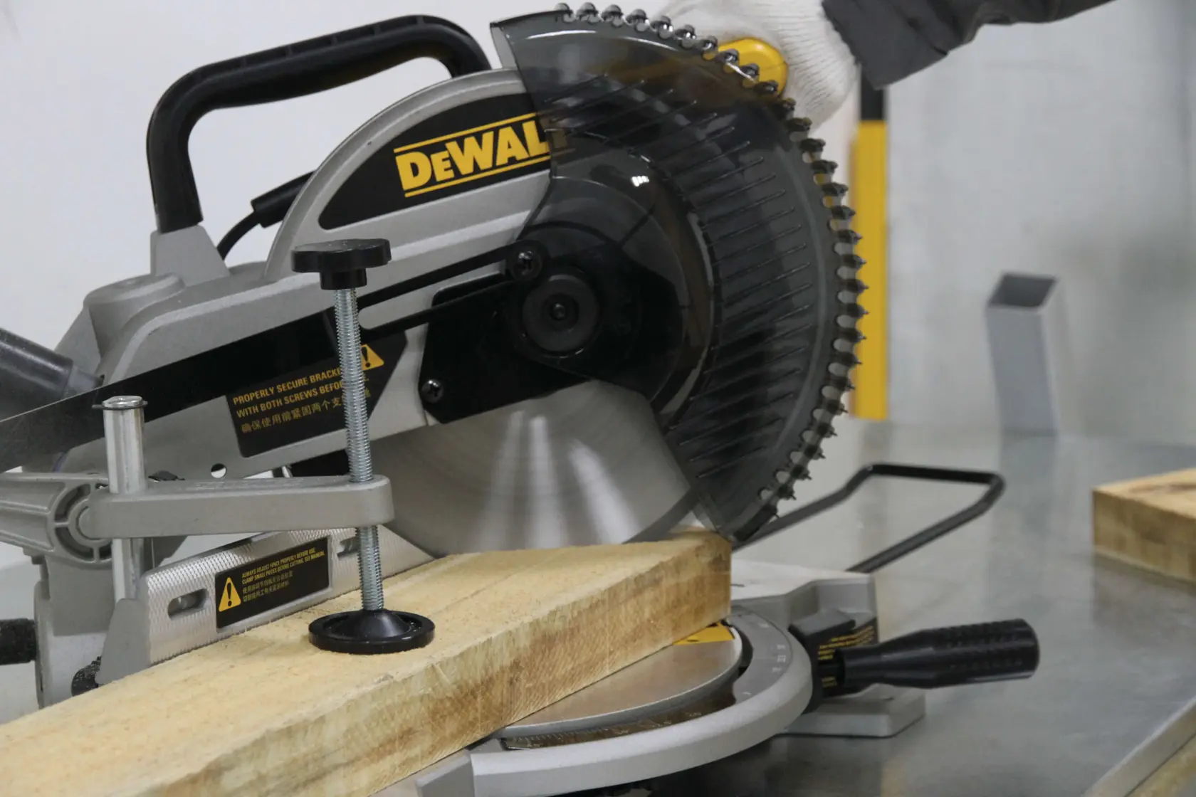 Una sierra de inglete DEWALT se utiliza para cortar un trozo de madera sobre una mesa de trabajo de metal. Una persona con un guante está operando la sierra.