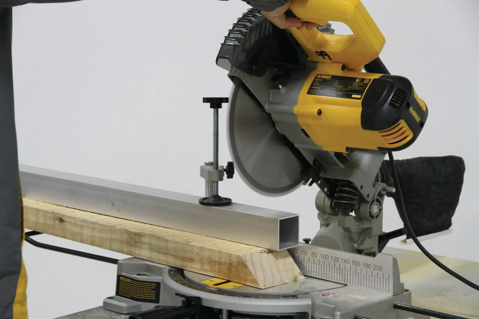Una sierra ingletadora amarilla DEWALT utilizada para cortar una pieza de metal y una de madera, aseguradas firmemente en la base de la sierra.