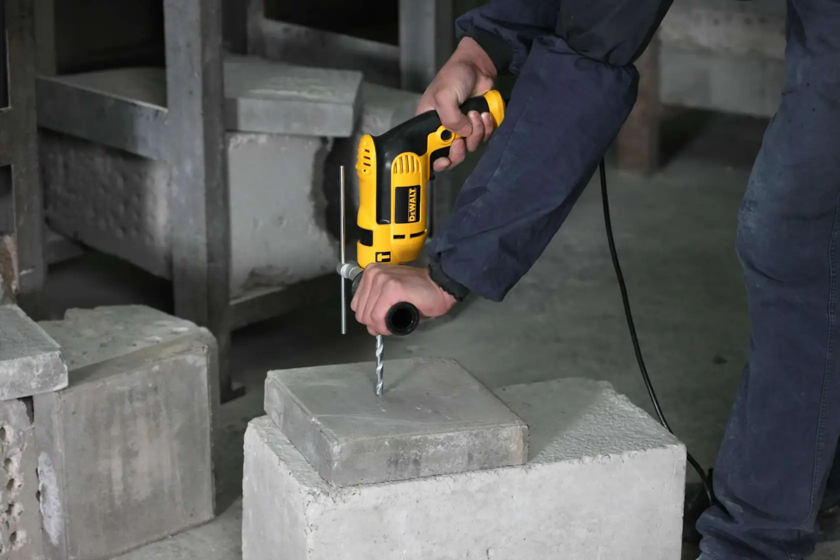 Una persona utiliza un taladro eléctrico amarillo DEWALT para perforar un bloque de concreto. La persona sostiene el taladro con ambas manos para mayor estabilidad en un entorno industrial.