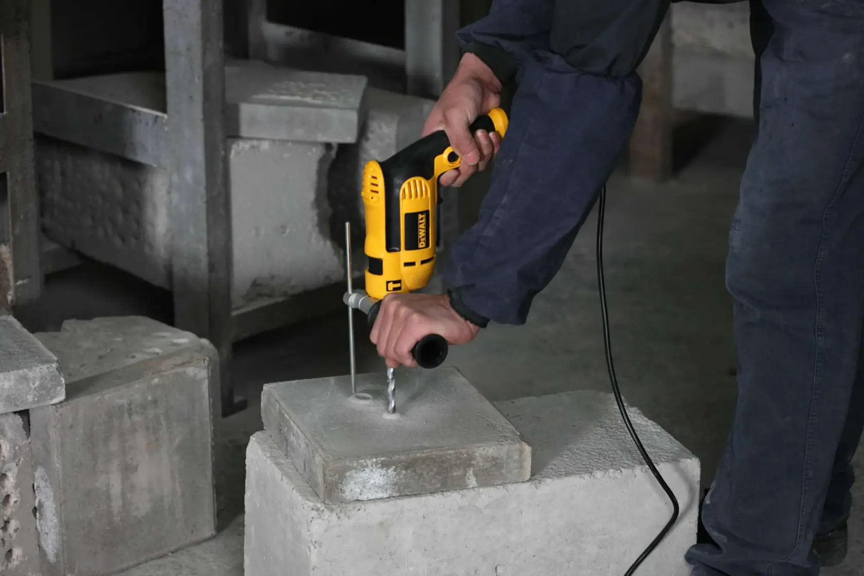 Una persona está utilizando un taladro eléctrico amarillo DEWALT para perforar un bloque de concreto en un entorno industrial.