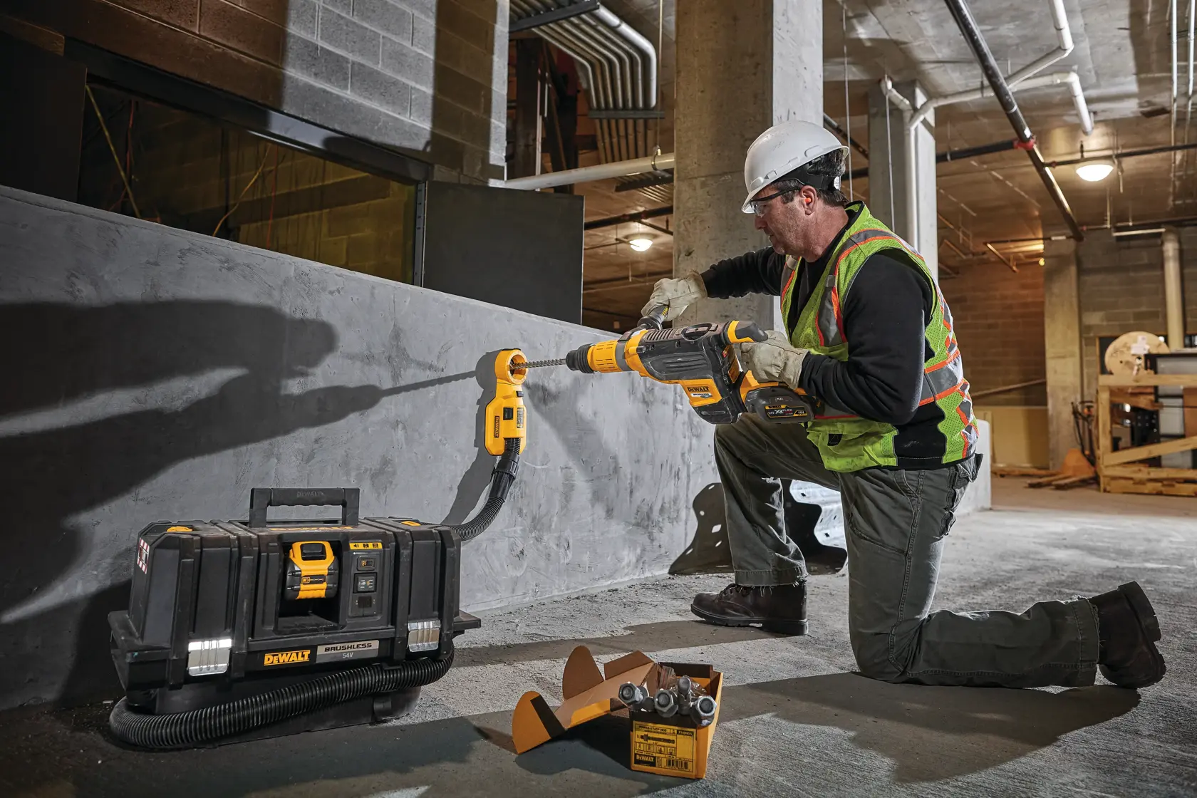 Una persona con equipo de seguridad opera un sistema de extracción de polvo DEWALT DWH050 conectado a un martillo rotativo, trabajando en una pared de concreto en un sitio de construcción. Una caja de herramientas DEWALT y accesorios de taladro están visibles en el suelo.