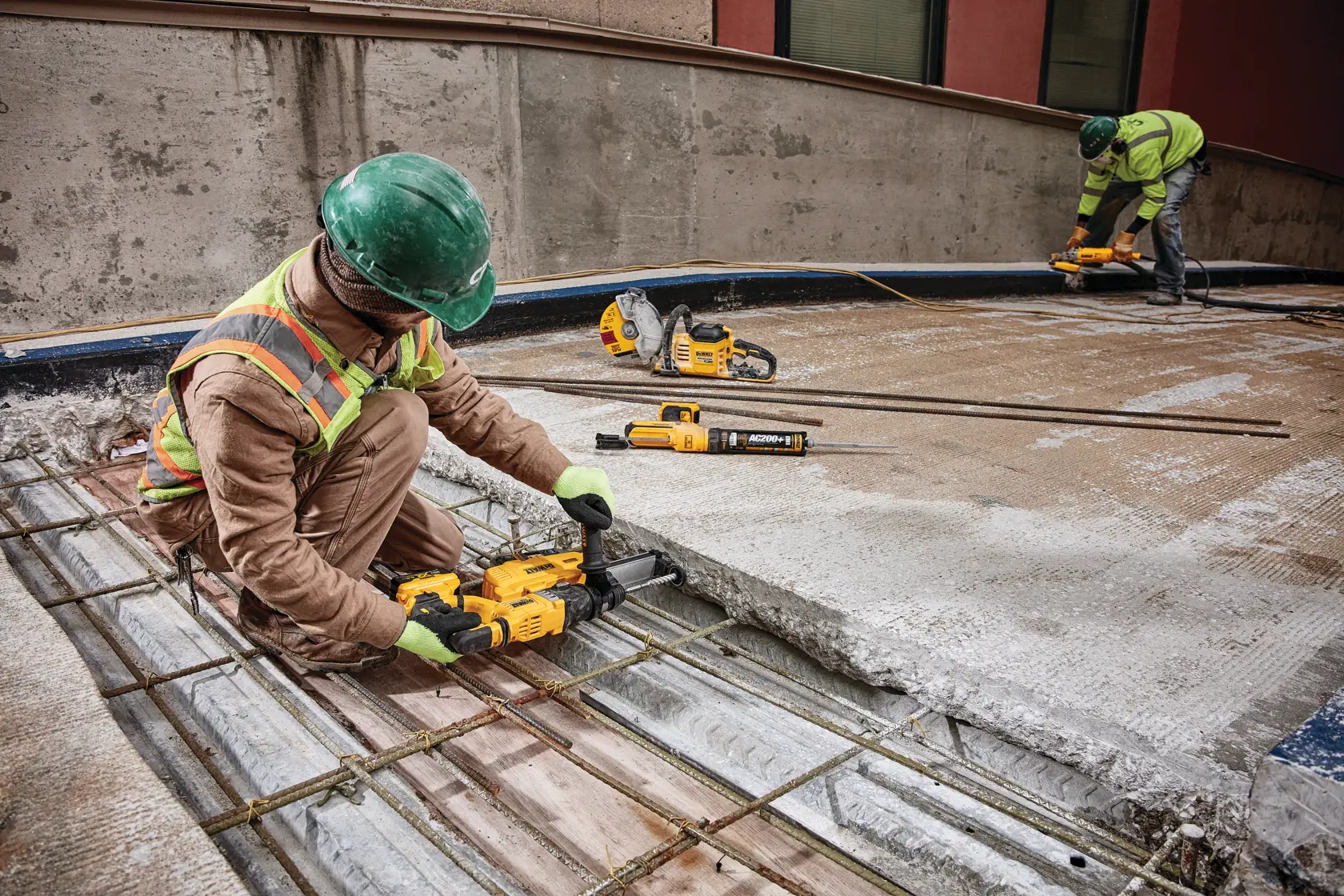 Dos trabajadores de la construcción con equipo de seguridad están usando herramientas DEWALT para trabajar sobre una superficie de concreto con barras de refuerzo de acero expuestas. Las herramientas y el equipo están ubicados alrededor del área de trabajo.