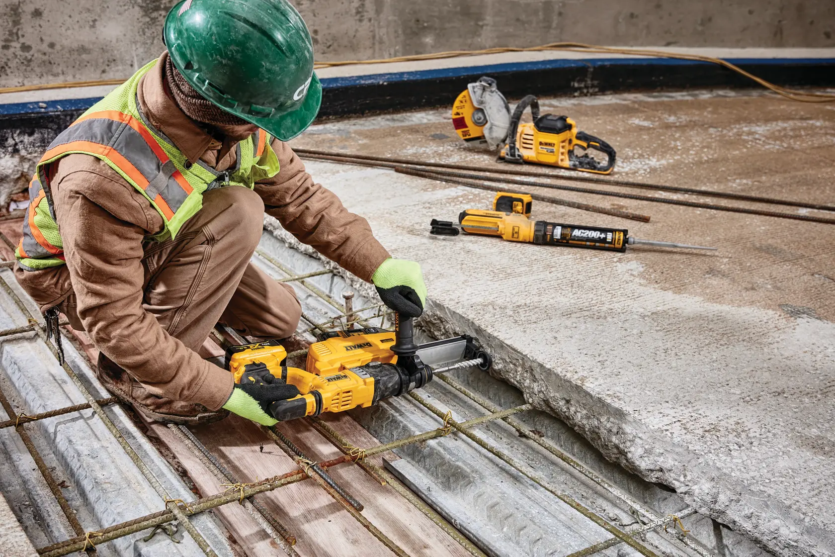 Un trabajador de construcción con equipo de seguridad y un casco verde utiliza una herramienta eléctrica amarilla DEWALT para perforar una losa de concreto. Más herramientas DEWALT, incluyendo una sierra y un martillo rotatorio, se ven en el suelo, todas destinadas a trabajos pesados de construcción.