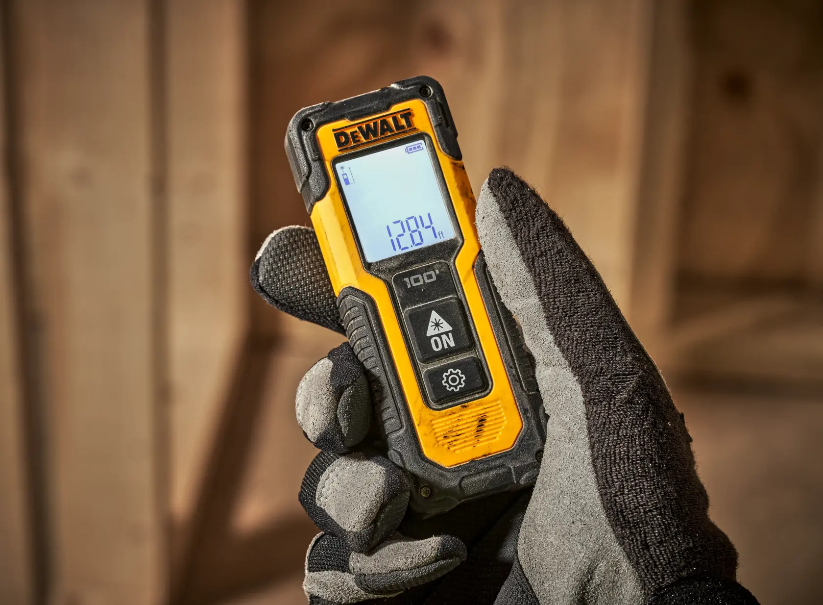 Una mano enguantada sostiene un medidor de distancia láser DEWALT con una pantalla digital que muestra lecturas de medición, en un entorno de construcción con vigas de madera al fondo.