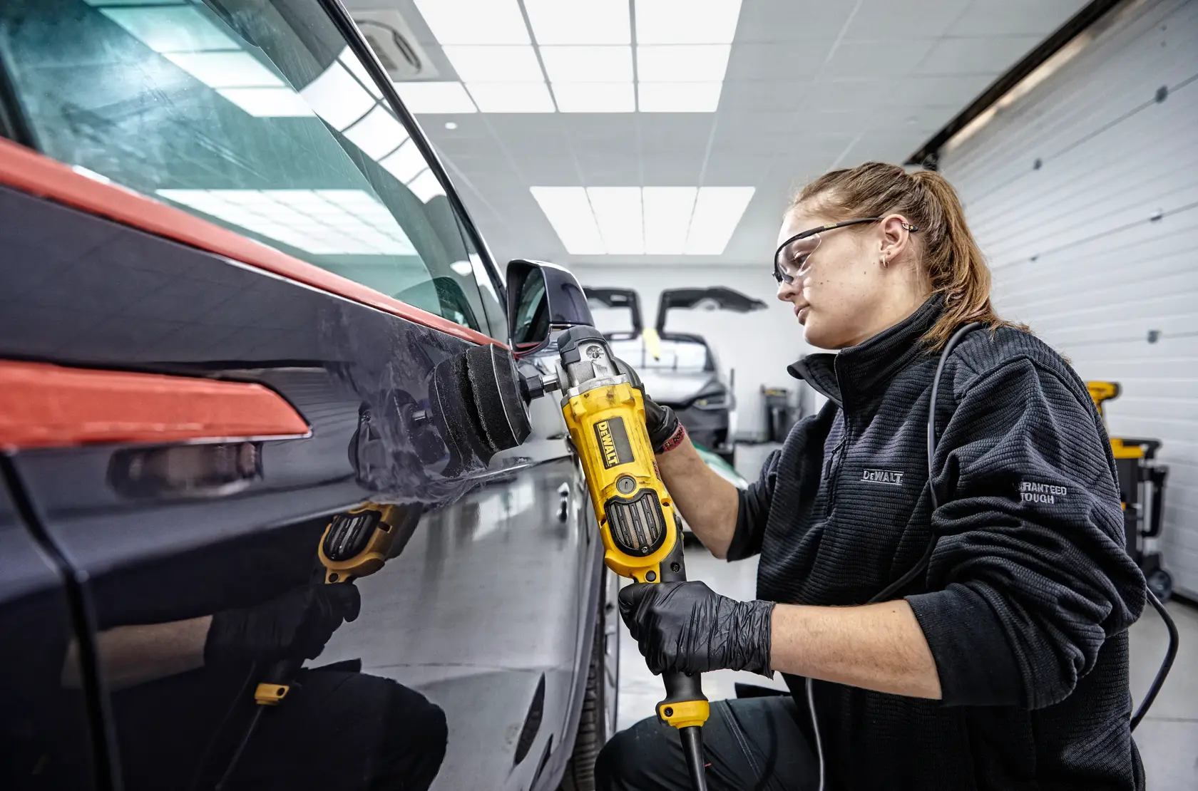 Persona pule puerta de coche usando herramienta amarilla DEWALT DWP849X en taller.