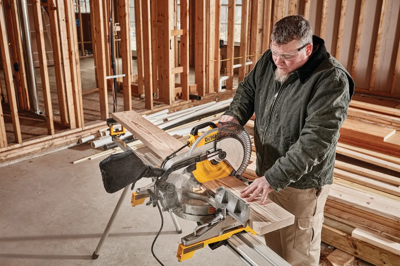 Una persona utiliza una sierra de inglete DEWALT DWS715 para cortar una tabla de madera en un sitio de construcción con estructuras de madera sin terminar y pilas de madera al fondo.