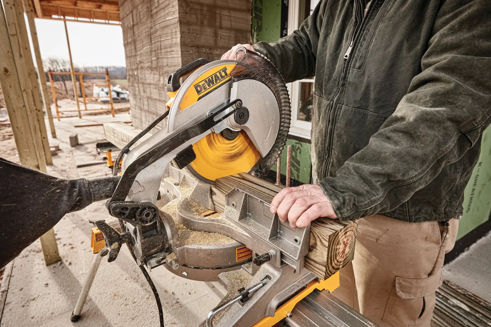 Una persona usando una sierra ingletadora DEWALT para cortar un trozo de madera al aire libre, con materiales de construcción y estructura visibles en el fondo.