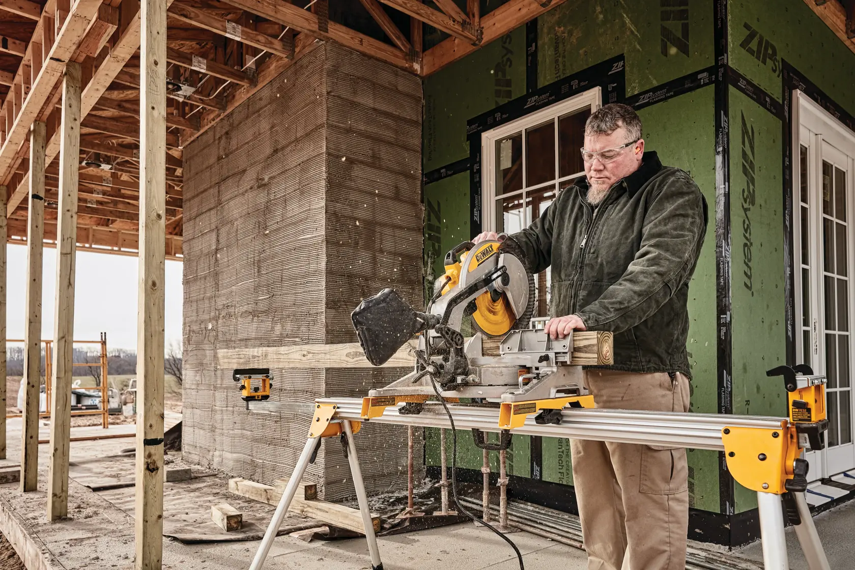 Una persona utiliza una sierra ingletadora DEWALT DWS715 para cortar un trozo de madera en un sitio de construcción. La sierra está montada en un soporte y hay una estructura de edificio sin terminar al fondo.