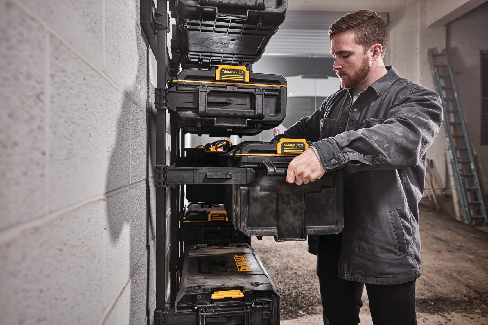Una persona organiza cajas de almacenamiento DEWALT en un estante montado en la pared de un taller o garaje. El rostro de la persona está difuminado y lleva una chaqueta de trabajo oscura.