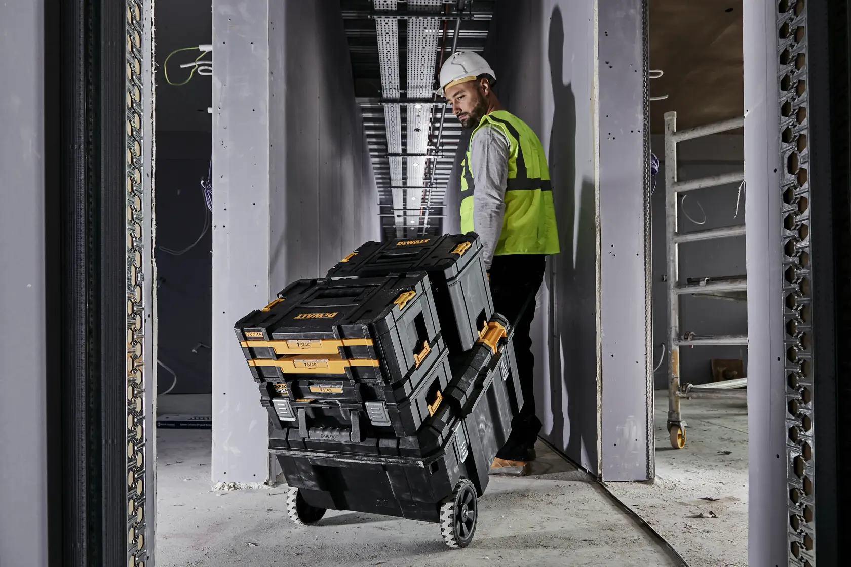 Trabajador lleva cajas de herramientas DEWALT apiladas con ruedas por un pasillo estrecho en obra.