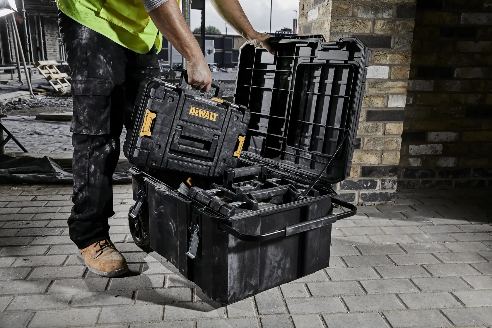 Persona coloca caja de herramientas DEWALT en un gran baúl abierto en obra con pared de ladrillos.
