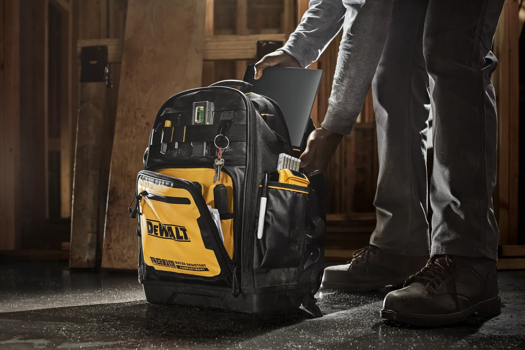Una mochila de herramientas DEWALT con varios compartimentos y herramientas organizadas, utilizada en un sitio de construcción. Una persona coloca una tableta en la mochila.
