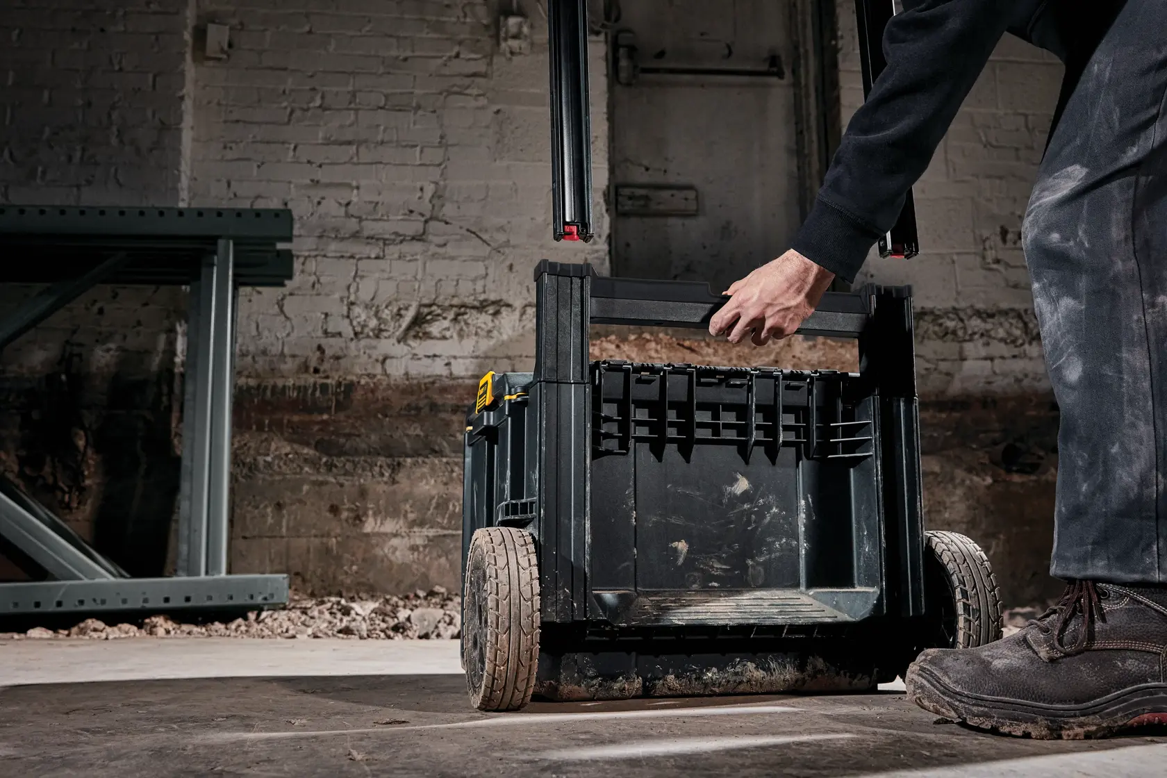 Una persona está manipulando una caja de almacenamiento portátil para herramientas DEWALT DWST60436 con ruedas en un sitio de construcción. La caja es negra y resistente, adecuada para transportar herramientas y equipos.