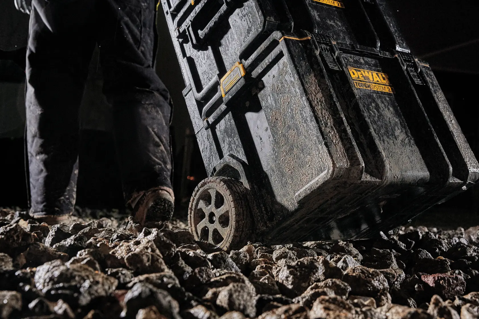 Vista de cerca de una caja de almacenamiento de herramientas con ruedas DEWALT ToughSystem siendo arrastrada sobre un terreno rocoso. La caja y las ruedas están cubiertas de barro, destacando la durabilidad y uso resistente.