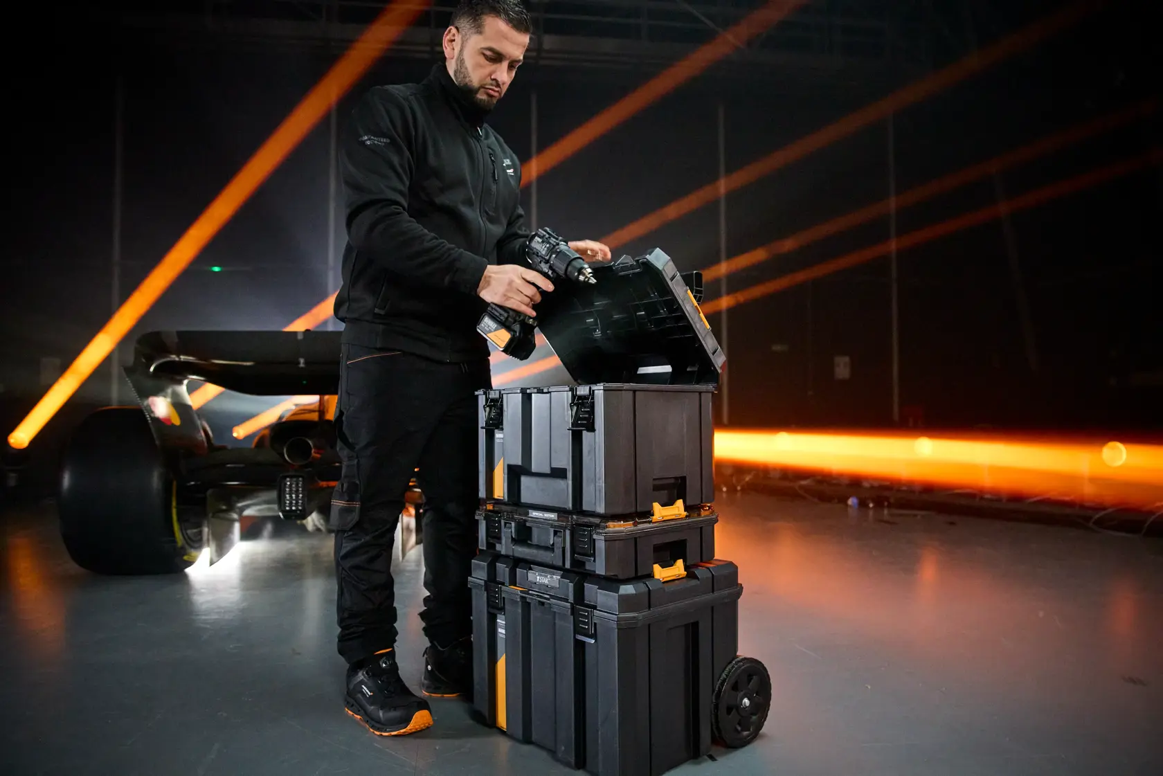 Persona usando apilado de cajas DEWALT con ruedas en ambiente industrial, junto a un auto deportivo iluminado.